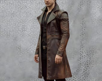 May include: A brown leather trench coat with a notched collar and a belt. The coat has a long, flowing design and is worn over a black vest and shirt. The coat's color is a mix of brown and bronze, with a slight sheen.