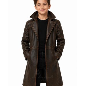 May include: A dark brown, distressed-finish leather trench coat featuring a wide lapel collar, long sleeves, and two front pockets. The coat is worn over a plain black t-shirt and black trousers, creating a layered look with a structured silhouette.