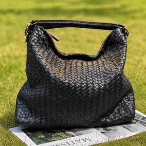 Puede incluir: Un bolso hobo de cuero trenzado negro con una silueta holgada, un asa superior única sujeta con anillos de metal dorado y un cierre de cremallera. El bolso descansa sobre una superficie rectangular blanca con el texto MATISSE impreso en negro.