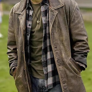 Op de afbeelding: Een lange bruine leren jas met een kraag en een versleten uitstraling. De persoon draagt een geruit overhemd, een groen shirt en een blauwe jeans. Een gouden hanger hangt aan een ketting.