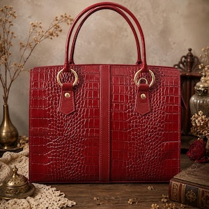 May include: A red leather tote bag with a crocodile texture. The bag has two top handles and gold-coloured metal rings. The bag is rectangular with a vertical seam down the centre. The background includes dried flowers and antique decor.