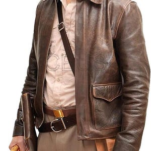 May include: A brown leather jacket with a collared neckline and two front pockets. The jacket has a distressed appearance and is worn over a light-coloured shirt. A brown belt with a gold buckle and a brown shoulder bag are also visible.