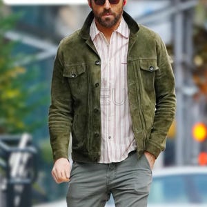 Op de afbeelding: Olijfgroene suède jas met knoopsluitingen en twee borstzakken. De jas wordt gedragen over een gestreept overhemd. De persoon draagt ook een grijze broek en een zonnebril.