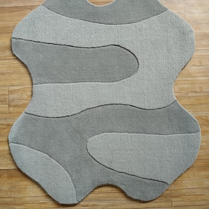 May include: A gray, abstract-shaped rug with a unique design. The rug features a layered pattern of light and medium gray sections, creating a textured appearance. The rug's edges have a wavy, organic shape, adding to its modern aesthetic.