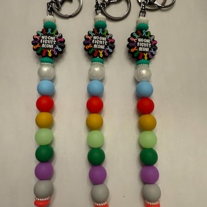 Könnte beinhalten: Drei Schlüsselanhänger mit einem Regenbogen aus Perlen. Jeder Schlüsselanhänger hat einen schwarzen Anhänger mit den Worten "NO ONE FIGHTS ALONE" und farbigen Awareness-Bändern. Die Schlüsselanhänger haben silberne Schlüsselringe und eine weiße Kordel.