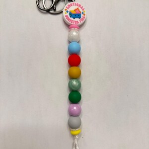 May include: A keychain with a round charm that reads "EMOTIONAL DUMPSTER FIRE" with a graphic of a dumpster on fire. The keychain has a series of colorful beads in a gradient pattern, including pink, white, blue, red, yellow, green, and gray.