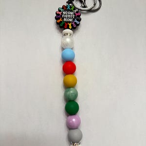 Può includere: Un portachiavi con un anello argentato e un design a perline. La parte superiore presenta un ciondolo nero con la scritta "NO ONE FIGHTS ALONE" circondata da nastri colorati. Sotto ci sono perline di vari colori: bianco, blu, rosso, giallo, verde, rosa e grigio.