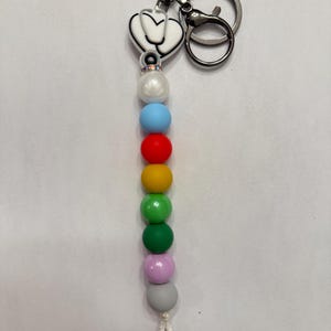 Puede incluir: Un llavero con un dije de estetoscopio y una cadena de cuentas de colores. El dije es blanco con un contorno negro. Las cuentas son de colores del arcoíris, incluyendo azul, rojo, amarillo, verde, rosa y gris. El llavero tiene un cierre plateado.
