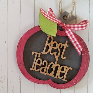 以下が含まれることがあります： 中央に金色の「Best Teacher」という文字が書かれた、ダークグレーの木製リンゴ型オーナメント。リンゴには赤いグリッターの縁取り、緑の葉、そして上部には赤と白のギンガムチェックのリボンとジュート麻の吊り下げ用ループが付いています。