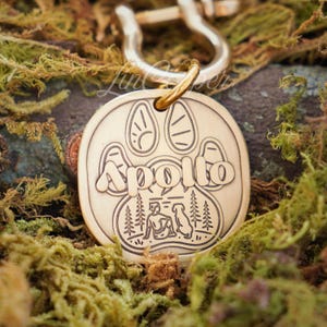 May include: A round, gold-colored pet tag with the name "Apollo" engraved in a paw print design. The tag features a small illustration of a person and a dog in a forest setting. A gold ring and silver clasp are attached.