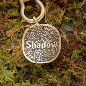 May include: A round, gold-colored pet tag with the name "Shadow" engraved in a bold font. The tag features a detailed floral pattern and is attached to a metal clasp. The background is a bed of green and brown moss.