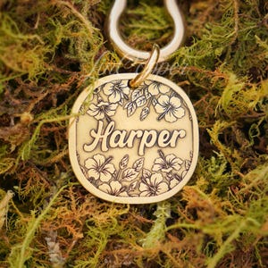 Könnte beinhalten: Ein runder, goldfarbener Hundeanhänger mit dem Namen "Harper" in eleganter Schrift graviert. Der Anhänger ist mit floralen Designs, darunter Hibiskusblüten, verziert. Oben ist eine goldfarbene Schlaufe zur einfachen Befestigung am Halsband angebracht. Der Anhänger liegt auf grünem und braunem Moos.