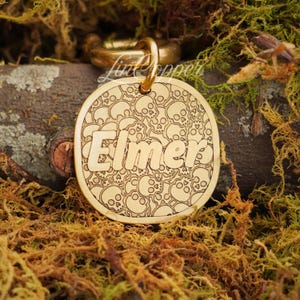 May include: A round gold-toned metal pet tag featuring an engraved pattern of multiple skulls across the surface. The name Elmer is prominently displayed in the center in a bold, sans-serif font. A gold-toned metal jump ring is attached at the top.