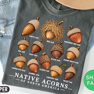 May include: A charcoal gray t-shirt featuring a botanical illustration of various acorns labeled White Oak, Bur Oak, Northern Red Oak, Pin Oak, Post Oak, Black Oak, Swamp White Oak, Chinkapin Oak, Scarlet Oak, Chestnut Oak, and Valley Oak with the text Native Acorns of North America.