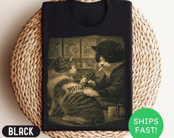 May include: Black t-shirt featuring a sepia-toned graphic of two cats. One cat is tattooing the word "MEOW" on the other cat's back. The tattoo artist cat is smoking a cigarette. The shirt has a round neckline.
