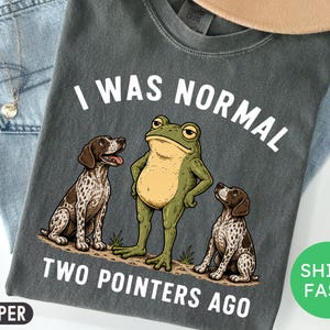 May include: A charcoal gray t-shirt featuring a graphic of a green frog standing between two brown and white spotted pointer dogs. The white text above reads I WAS NORMAL and the text below reads TWO POINTERS AGO. The shirt has a crew neckline.