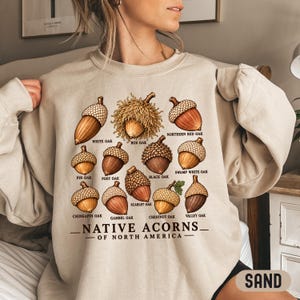 May include: A sand-colored crewneck sweatshirt featuring a graphic print of various acorns labeled White Oak, Bur Oak, Northern Red Oak, Pin Oak, Post Oak, Black Oak, Swamp White Oak, Chinkapin Oak, Gambel Oak, Scarlet Oak, Chestnut Oak, Valley Oak, and Native Acorns of North America.