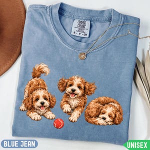 May include: A blue jean colored t-shirt featuring a graphic of three brown and white curly-haired dogs. One dog is in a play bow position, one is running, and one is sleeping. A small red ball is positioned near the dogs. The shirt has a Comfort Colors label.