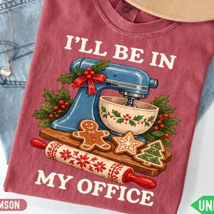 May include: A crimson t-shirt featuring a blue stand mixer, a mixing bowl, a rolling pin, and gingerbread cookies. The text reads I'LL BE IN MY OFFICE. The design includes holly leaves and red berries, with a cream-colored bowl decorated with holly.