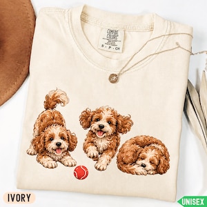 May include: An ivory-colored t-shirt featuring a graphic print of three brown and white curly-haired puppies. One puppy is in a play bow position, one is running, and one is sleeping next to a small red ball. The shirt has a Comfort Colors brand label.