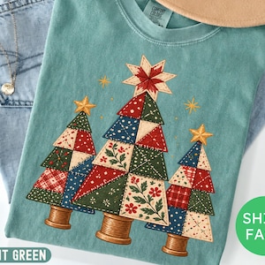 May include: A light green t-shirt featuring a graphic of three Christmas trees designed with a patchwork quilt pattern in red, green, and blue. Each tree is topped with a yellow star and sits on a wooden spool base. The shirt has a crew neckline.