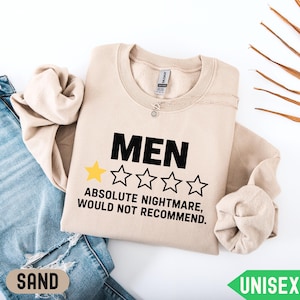 May include: A sand-colored crewneck sweatshirt featuring the black text MEN above a graphic of one yellow star and four empty star outlines. Below the stars, the black text reads ABSOLUTE NIGHTMARE, WOULD NOT RECOMMEND. The garment has long sleeves.