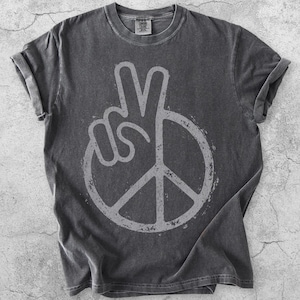 Peut inclure: Un t-shirt gris anthracite à manches courtes avec un motif gris clair vieilli représentant un signe de paix combiné à une main faisant un geste de paix. Le t-shirt possède un col rond, des manches retroussées et une étiquette de marque visible.
