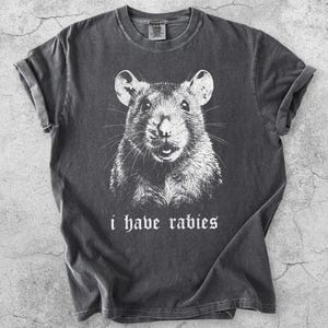 May include: A charcoal grey short-sleeve t-shirt featuring a white graphic illustration of a rat's face centred on the chest. Below the rat, the text i have rabies is printed in a white gothic-style font. The shirt has a crew neckline.