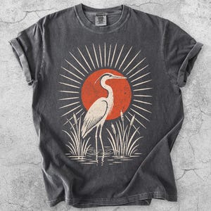 Pu&ograve; includere: Una t-shirt grigio antracite con una stampa grafica di un airone bianco in piedi tra i canneti davanti a un grande sole arancione con linee bianche radianti. La maglietta ha un girocollo, maniche corte e un'etichetta visibile sul colletto.