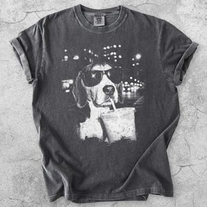 May include: A charcoal grey short-sleeve t-shirt featuring a black and white graphic print of a dog wearing dark sunglasses and drinking from a plastic cup with a straw. The shirt has a crew neckline and a vintage-style washed fabric finish.
