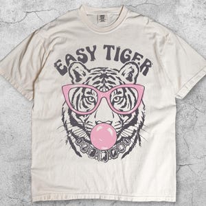 May include: An ivory short-sleeve t-shirt featuring a graphic of a tiger wearing pink glasses and a jewelled necklace while blowing a pink bubble. The text "EASY TIGER" is printed in a dark grey arched font above the tiger illustration.