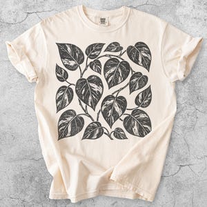 Op de afbeelding: Een ivoorkleurig T-shirt met korte mouwen en een centrale zwarte grafische print van meerdere hartvormige bladeren met een verweerde, getextureerde uitstraling. Het shirt heeft een ronde hals en een standaard pasvorm, met het woord Ivory onderaan.
