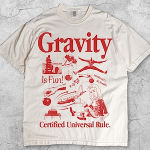 Könnte beinhalten: Elfenbeinfarbenes T-Shirt mit rotem Grafikdesign. Das Design enthält das Wort "Gravity" über verschiedenen Illustrationen und den Text "Is Fun!" und "Certified Universal Rule."
