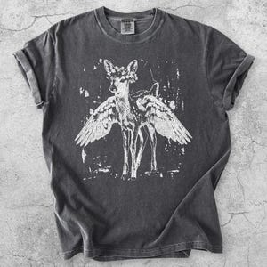 Op de afbeelding: Donkergrijze t-shirt met een witte afbeelding van een hert met vleugels en een bloemenkrans. De tee heeft een vintage, distressed look met opgerolde mouwen. Het woord "Pepper" staat linksonder.