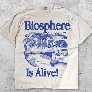 以下が含まれることがあります： アイボリー色のTシャツ。青色のグラフィックデザインで、「Biosphere」の文字と風景のイラストが描かれています。「Is Alive!」の文字も含まれています。「Where air, water, and land meet」のテキストも含まれています。