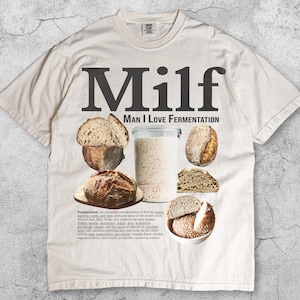 Può includere: Una t-shirt color avorio con il testo "Milf Man I Love Fermentation" in grandi lettere grigio scuro. La grafica include immagini di varie pagnotte di lievito naturale e un barattolo di vetro di lievito, accompagnate da un blocco di testo.
