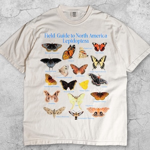 Puede incluir: Camiseta de color marfil con un gráfico detallado de varias mariposas norteamericanas y sus nombres. El texto "Field Guide to North America Lepidoptera" está impreso en azul. La camiseta está hecha de un material suave.