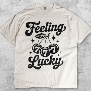 May include: Ivory-colored t-shirt with the words "Feeling Lucky" in a bold, black script. The design features a cluster of cherries with the number "7" inside each cherry. The shirt is a casual, unisex style.