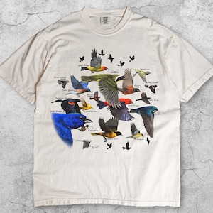 May include: Ivory-colored t-shirt featuring a detailed illustration of various bird species in flight. The design includes colorful birds with their names labeled. The shirt is made of a soft material.