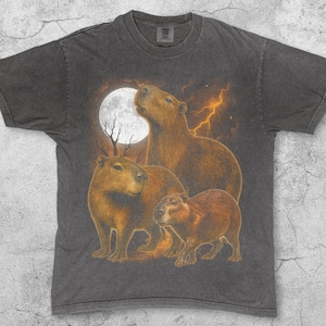 Op de afbeelding: Donkergrijs t-shirt met een capybarafamilieontwerp onder een volle maan en bliksem. De capibara's zijn bruin en bruingeel. De grootste capibara kijkt naar de maan. Het woord "Pepper" staat linksonder.
