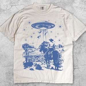 May include: Ivory-colored t-shirt with a blue graphic of a cowboy on a horse being abducted by a UFO. The design includes a desert landscape, stars, and the word "Ivory" at the bottom.