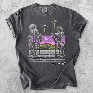 Könnte beinhalten: Dunkelgraues T-Shirt mit einer Grafik einer Wüstenlandschaft unter einem lila und rosa Himmel, mit UFOs und dem Text "Area 51, NV". Das T-Shirt hat ein Gittermuster und Joshua-Bäume.
