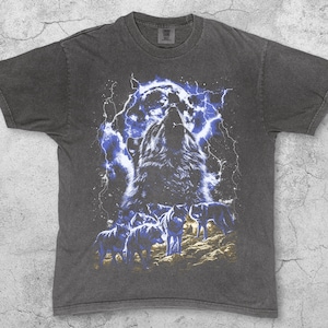 Puede incluir: Camiseta color pimienta con un gr&aacute;fico de un lobo aullando sobre un fondo de tormenta el&eacute;ctrica azul. Varios lobos se representan debajo. La palabra "Pepper" est&aacute; impresa en la parte inferior de la camiseta.