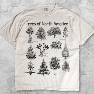 May include: Ivory-colored t-shirt featuring a graphic of various North American trees, including Giant Sequoia and Joshua Tree. The text "Trees of North America" is printed above the illustrations. The shirt is made of a soft material.