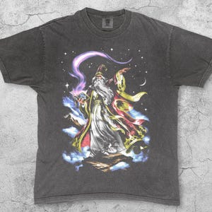 Könnte beinhalten: Pfefferfarbenes T-Shirt mit einer detaillierten Illustration eines Zauberers mit langem, weißem Bart, der ein leuchtendes Buch und einen Stab hält. Der Zauberer ist von Sternen, Wolken und einem Halbmond umgeben. Das Wort "Pepper" ist unten aufgedruckt.