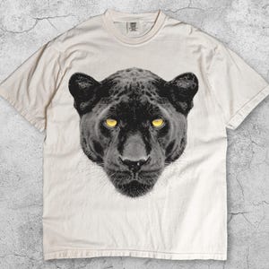 May include: Ivory-colored t-shirt featuring a detailed black panther face with yellow eyes. The graphic is centered on the shirt. The word "Ivory" is printed at the bottom left. The shirt has a classic crew neck design.
