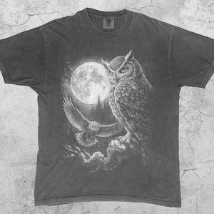 May include: Pepper-colored t-shirt featuring a detailed illustration of two owls under a full moon. One owl is perched, and the other is in flight. The design is in shades of gray and white.