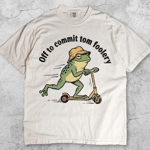 May include: Ivory t-shirt with a cartoon frog on a scooter, wearing a bucket hat. The text "Off to commit tom foolery" is above the frog. The shirt has a crew neck and short sleeves. The word "Ivory" is at the bottom.