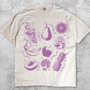 May include: Ivory-colored t-shirt featuring a purple fruit illustration. The design includes grapes, a pear, a sliced orange, bananas, an apple, a raspberry, a peach, a pomegranate, and a pineapple. The word "Ivory" is printed at the bottom.