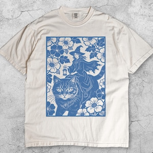 Op de afbeelding: Ivoor kleurig t-shirt met een blauwe illustratie van een heks die op een kat rijdt, omringd door bloemen. De heks houdt een lantaarn vast. Het woord "Ivory" staat onderaan het shirt.